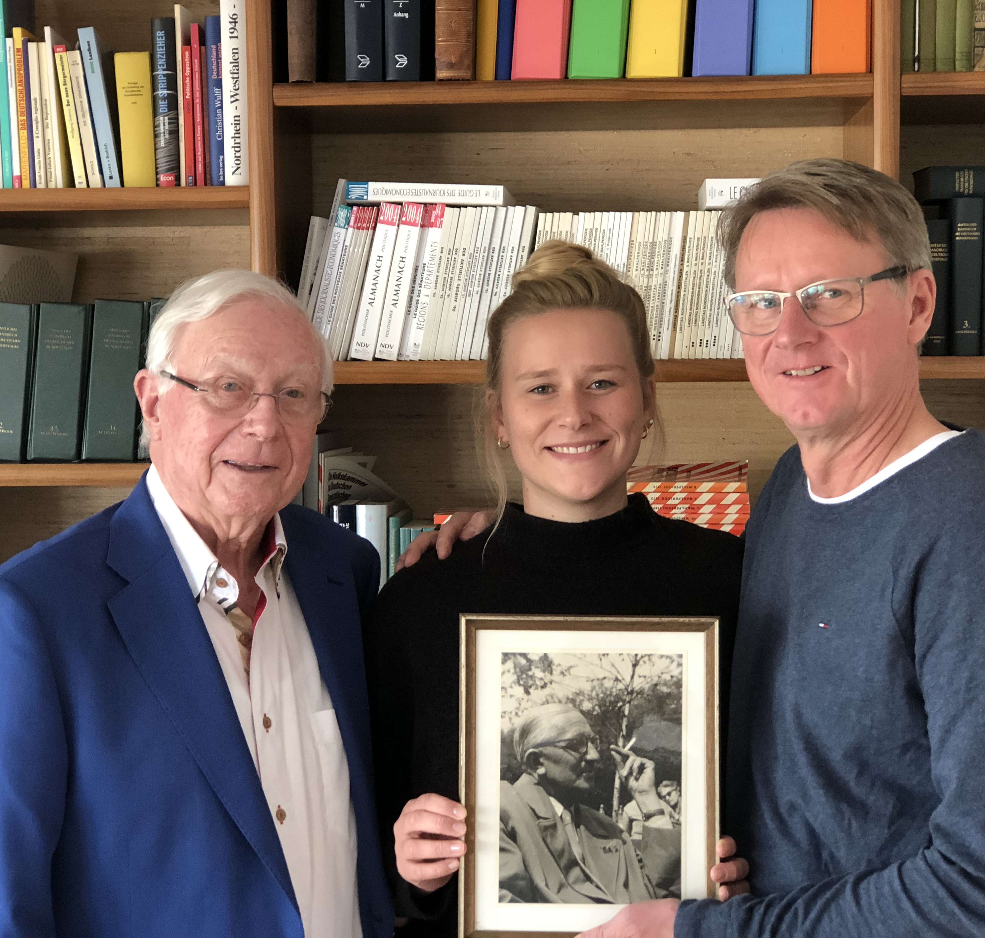 Im Bild vor einer Bücherwand: Klaus-J. Holzapfel, Katrin Holzapfel und Andreas Holzapfel, die gemeinsam ein Foto von Adolf Holzapfel in den Händen halten.